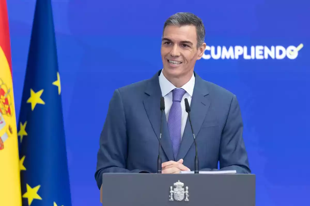 Hombre de traje azul hablando en un podio con banderas de España y la Unión Europea al fondo y un fondo azul con la palabra cumpliendo parcialmente visible Hombre de traje azul hablando en un podio con banderas de España y la Unión Europea al fondo y un fondo azul con la palabra cumpliendo parcialmente visible