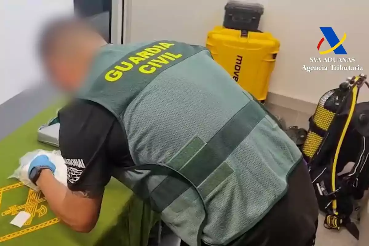 Agente de la Guardia Civil revisando un paquete sobre una mesa verde junto a equipo de buceo y maletas en una sala con el logo de la Agencia Tributaria en la pared