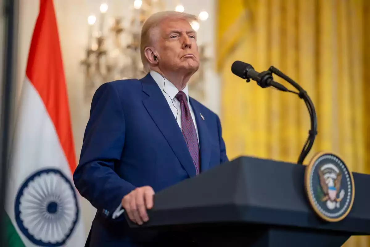 Hombre de cabello rubio con traje azul hablando en un podio presidencial junto a una bandera de la India