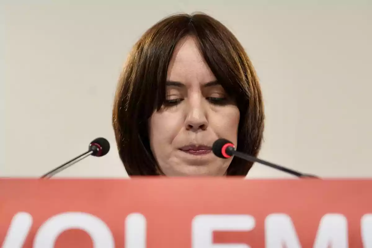 Mujer de cabello castaño corto mirando hacia abajo detrás de un podio rojo con dos micrófonos frente a ella