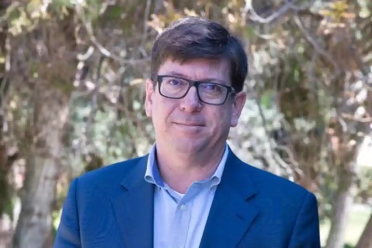 Hombre de cabello corto y gafas con saco azul posando al aire libre frente a árboles desenfocados Hombre de cabello corto y gafas con saco azul posando al aire libre frente a árboles desenfocados