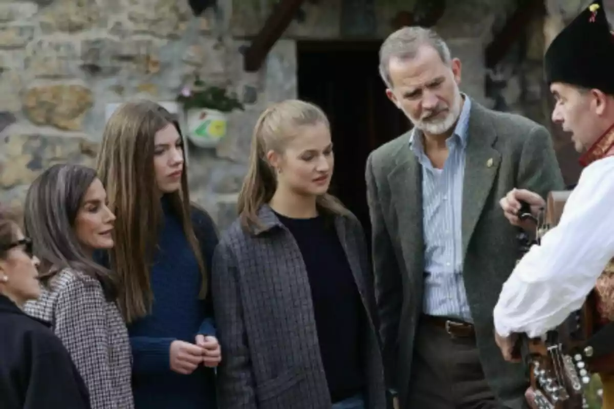 A group of people—who are the Spanish Royal Family—are watching closely as a man dressed in traditional clothing holds a musical instrument in front of a stone wall. A group of people—who are the Spanish Royal Family—are watching closely as a man dressed in traditional clothing holds a musical instrument in front of a stone wall.