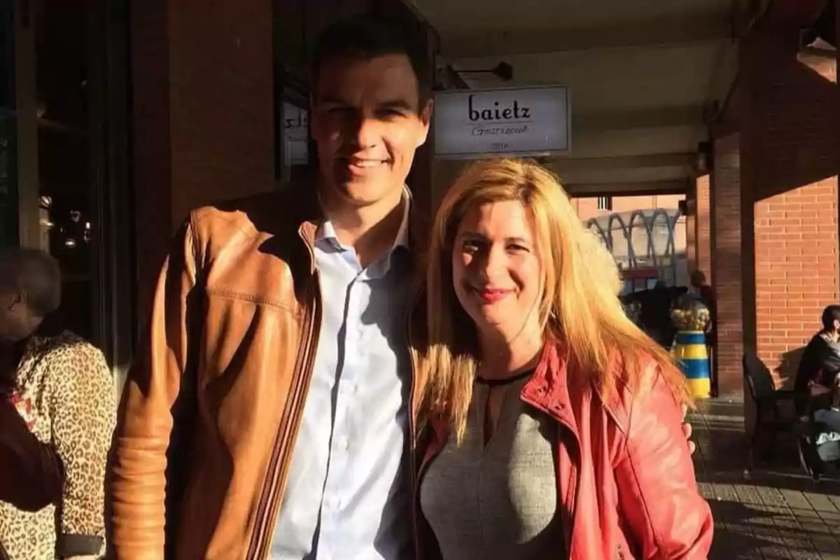 Dos personas sonríen y posan juntas al aire libre en una zona peatonal con luz de sol, ambas llevan chaquetas y al fondo se ven otras personas y un letrero que dice baietz. Dos personas sonríen y posan juntas al aire libre en una zona peatonal con luz de sol, ambas llevan chaquetas y al fondo se ven otras personas y un letrero que dice baietz.