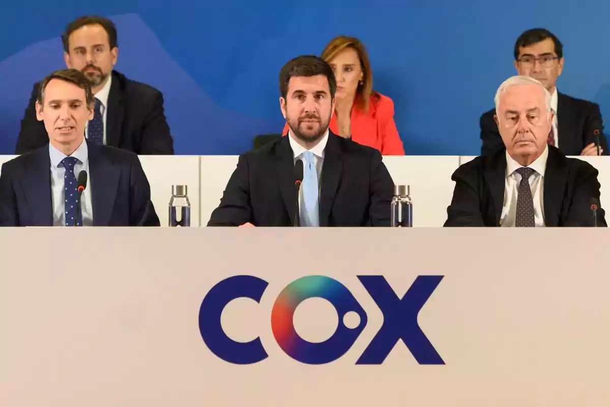 Tres hombres con traje sentados en una mesa de conferencia con el logo de COX al frente y tres personas más al fondo Tres hombres con traje sentados en una mesa de conferencia con el logo de COX al frente y tres personas más al fondo