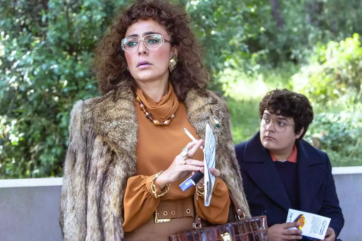 Mujer con abrigo de piel y gafas sostiene un cigarrillo y papeles mientras un joven con gafas la observa sentado en un entorno al aire libre con vegetación de fondo Mujer con abrigo de piel y gafas sostiene un cigarrillo y papeles mientras un joven con gafas la observa sentado en un entorno al aire libre con vegetación de fondo