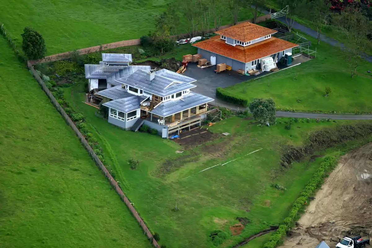 Vista aérea de una casa grande con techo gris y una construcción adicional con techo naranja, rodeadas de césped verde y un camino de entrada, en una zona rural Vista aérea de una casa grande con techo gris y una construcción adicional con techo naranja, rodeadas de césped verde y un camino de entrada, en una zona rural