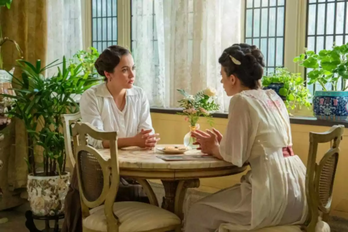 Dos mujeres vestidas con ropa de época conversan sentadas en una mesa redonda junto a una ventana rodeadas de plantas y luz natural Dos mujeres vestidas con ropa de época conversan sentadas en una mesa redonda junto a una ventana rodeadas de plantas y luz natural