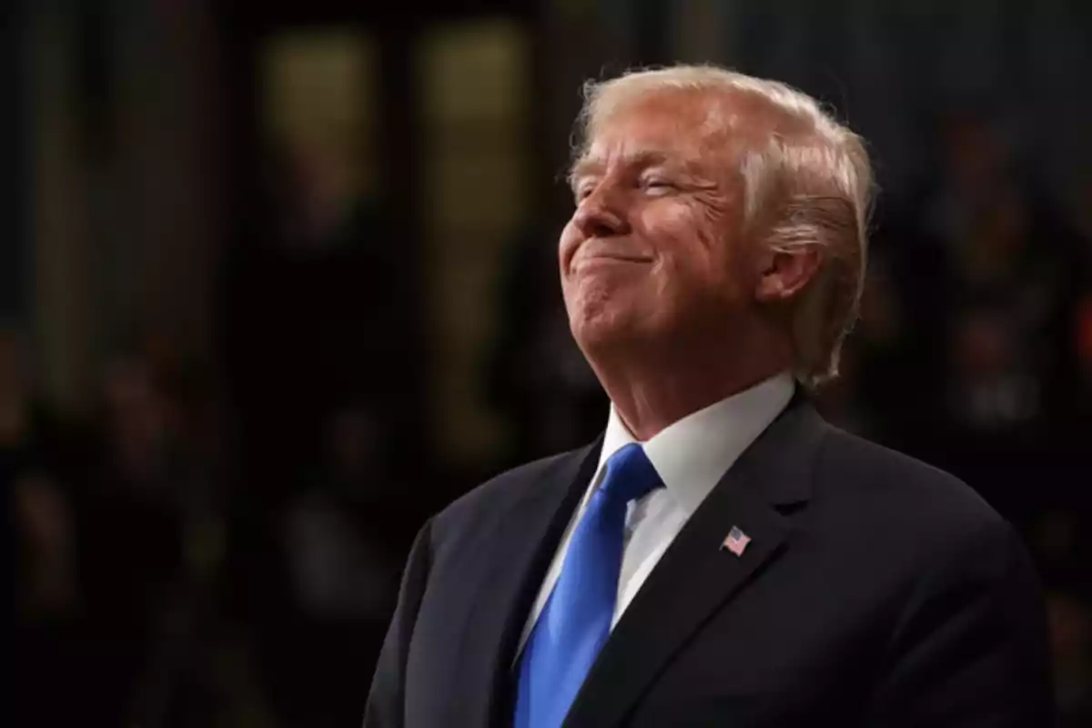 Hombre de cabello rubio con traje oscuro y corbata azul sonriendo con expresión de satisfacción en un entorno formal Hombre de cabello rubio con traje oscuro y corbata azul sonriendo con expresión de satisfacción en un entorno formal