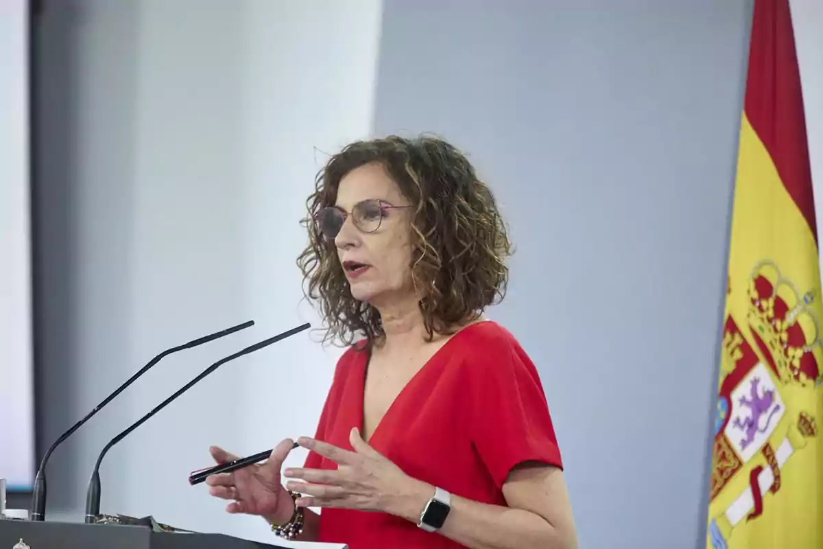 Mujer con gafas y vestido rojo hablando frente a micrófonos con una bandera de España al fondo Mujer con gafas y vestido rojo hablando frente a micrófonos con una bandera de España al fondo