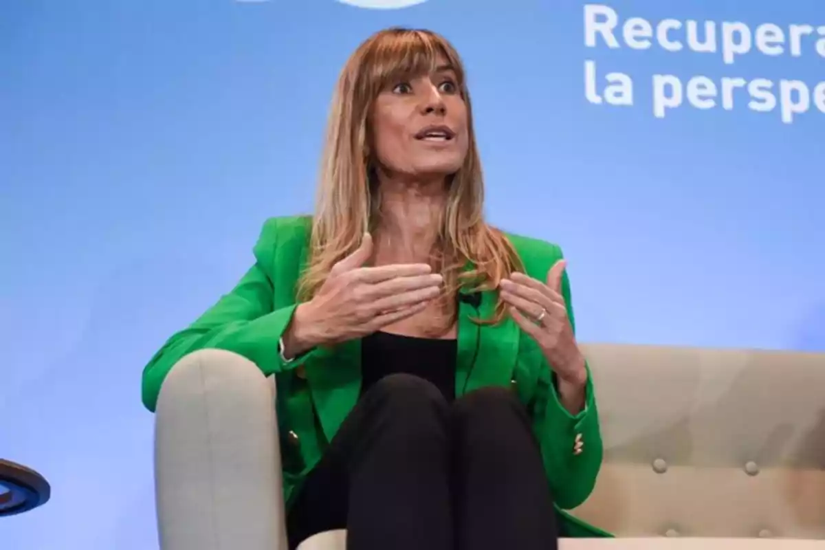 Mujer sentada en un sillón hablando y gesticulando con las manos, lleva un saco verde y fondo azul con texto parcial. Mujer sentada en un sillón hablando y gesticulando con las manos, lleva un saco verde y fondo azul con texto parcial.