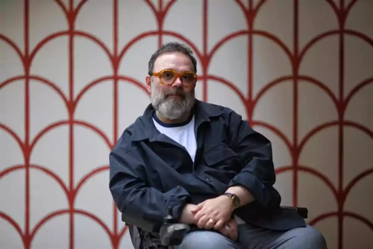 Hombre con barba y gafas sentado en una silla de ruedas frente a una pared con patrón geométrico rojo
