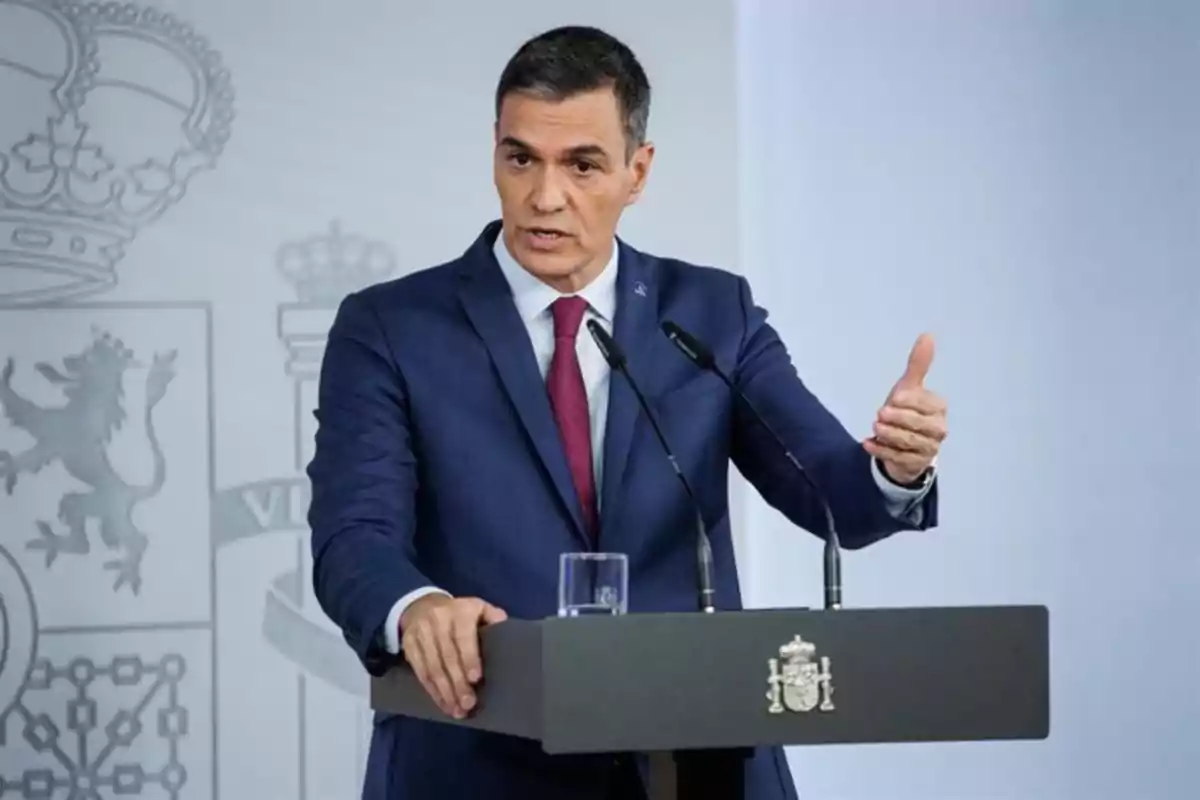 Hombre con traje azul y corbata roja hablando en una conferencia de prensa detrás de un atril con el escudo de España Hombre con traje azul y corbata roja hablando en una conferencia de prensa detrás de un atril con el escudo de España