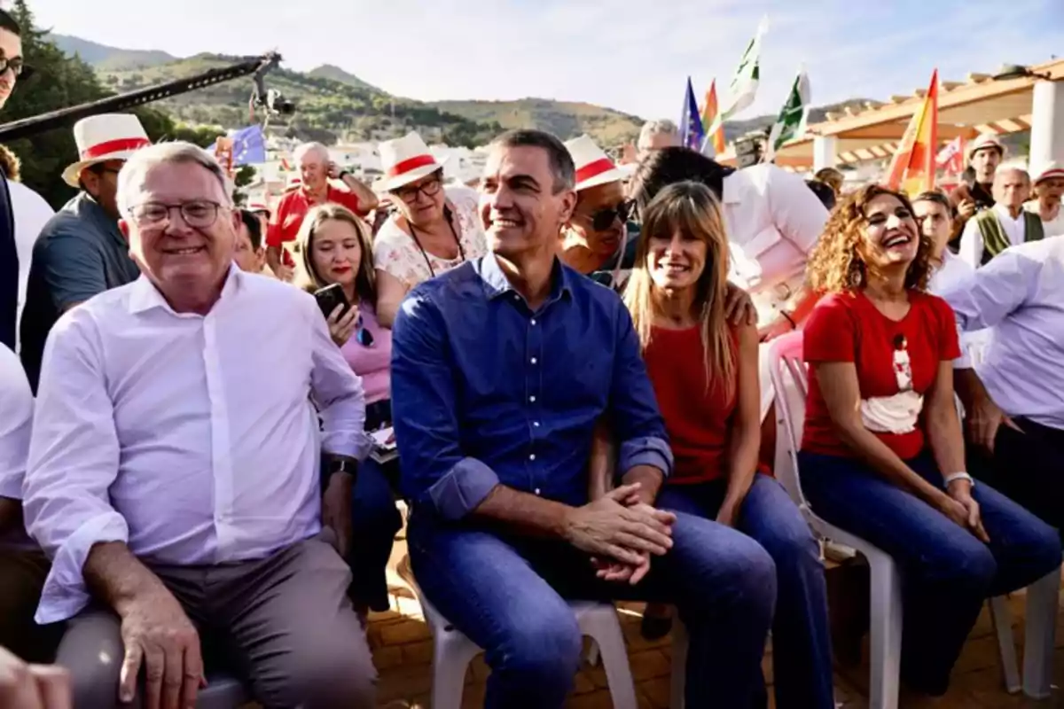 Grupo de personas sentadas al aire libre sonriendo y disfrutando de un evento festivo con banderas y montañas al fondo Grupo de personas sentadas al aire libre sonriendo y disfrutando de un evento festivo con banderas y montañas al fondo