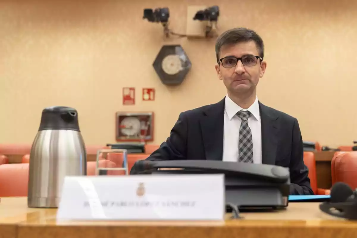 Hombre con gafas y traje sentado en una sala de reuniones con una cafetera de acero inoxidable y un vaso de agua sobre la mesa Hombre con gafas y traje sentado en una sala de reuniones con una cafetera de acero inoxidable y un vaso de agua sobre la mesa