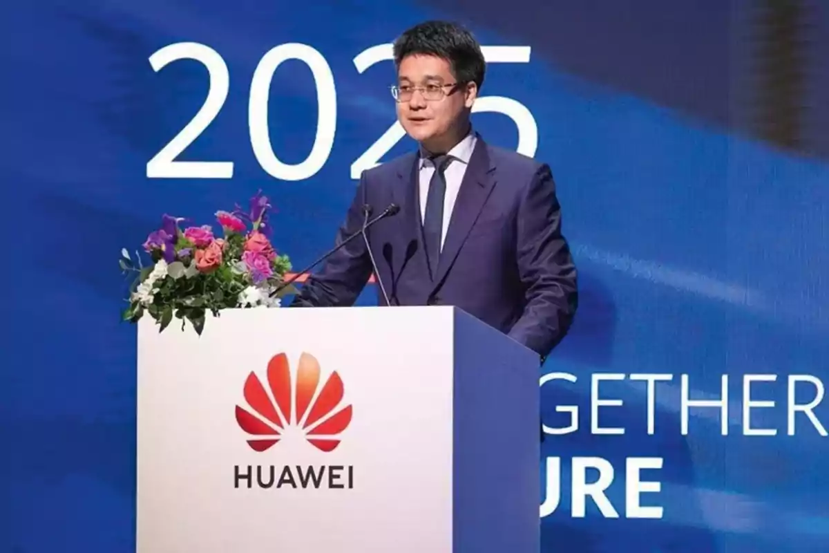 Hombre con gafas y traje azul hablando en un podio con el logo de Huawei y flores, fondo azul con el número 2025 y texto en inglés Hombre con gafas y traje azul hablando en un podio con el logo de Huawei y flores, fondo azul con el número 2025 y texto en inglés
