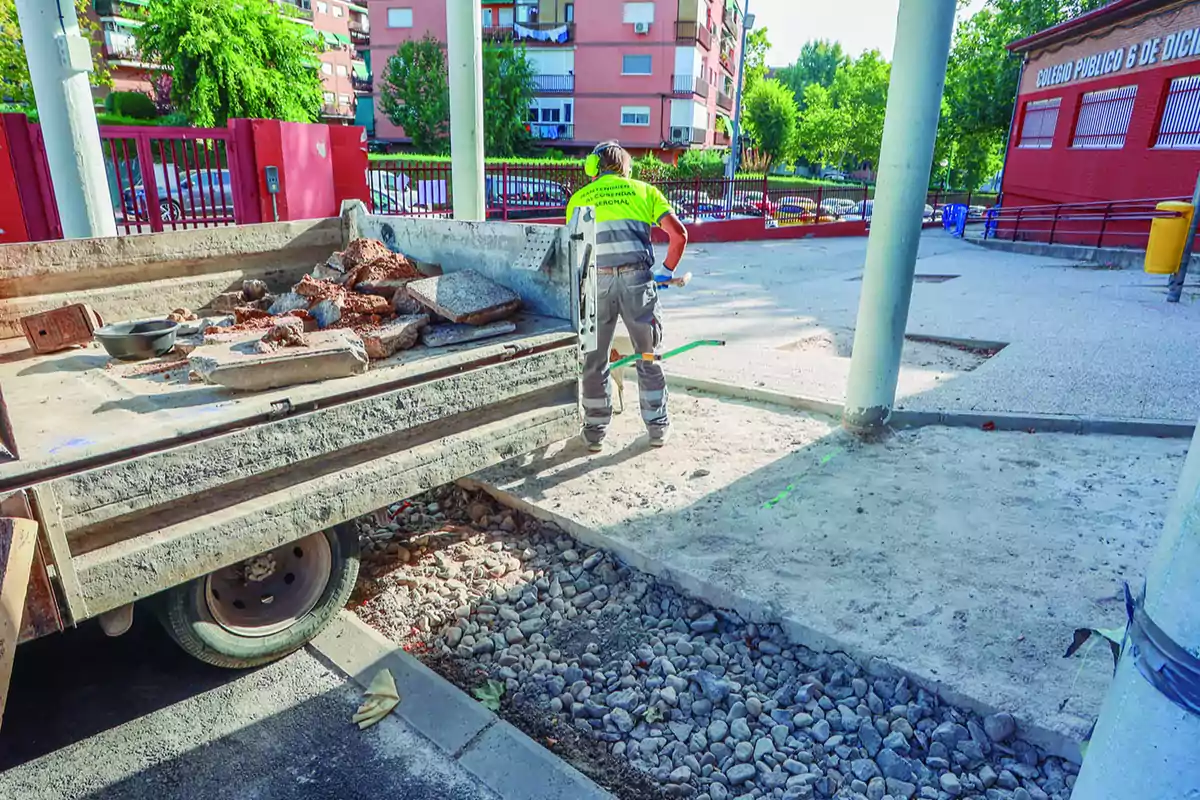 Trabajador de la construcción con chaleco reflectante realizando labores de mantenimiento en una zona exterior junto a un camión con escombros y un colegio al fondo Trabajador de la construcción con chaleco reflectante realizando labores de mantenimiento en una zona exterior junto a un camión con escombros y un colegio al fondo