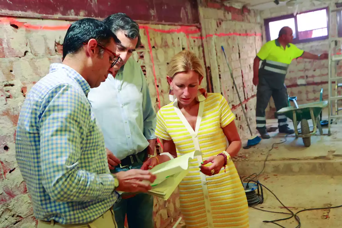 Tres personas revisan documentos en una obra en construcción mientras un trabajador está al fondo junto a una carretilla y herramientas