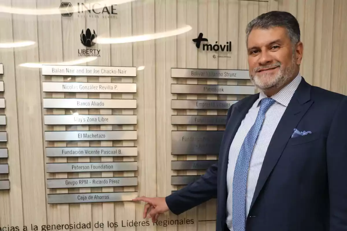 Hombre de traje sonriente señalando una placa en una pared con nombres de empresas y fundaciones en un entorno corporativo