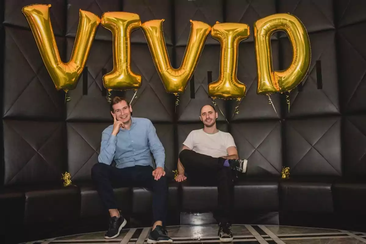 Dos hombres sentados en un sofá negro con globos dorados que forman la palabra VIVID detrás de ellos