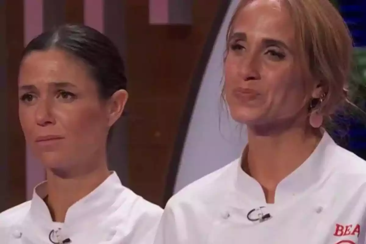 Gabriela y Bea con chaquetas de chef blancas muestran expresiones serias y emotivas en un set de competencia culinaria de MasterChef. Gabriela y Bea con chaquetas de chef blancas muestran expresiones serias y emotivas en un set de competencia culinaria de MasterChef.
