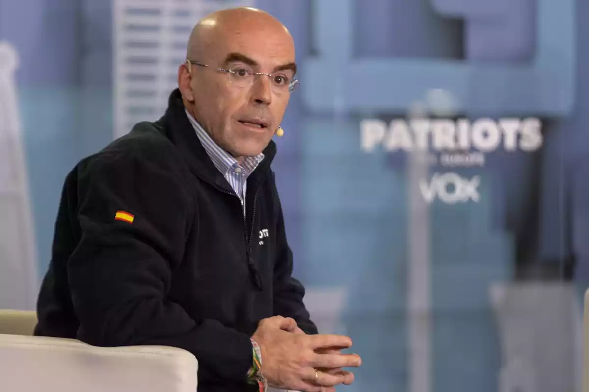 Hombre calvo con gafas y chaqueta oscura sentado en un sillón durante un evento con el logo de VOX y la palabra Patriots en el fondo