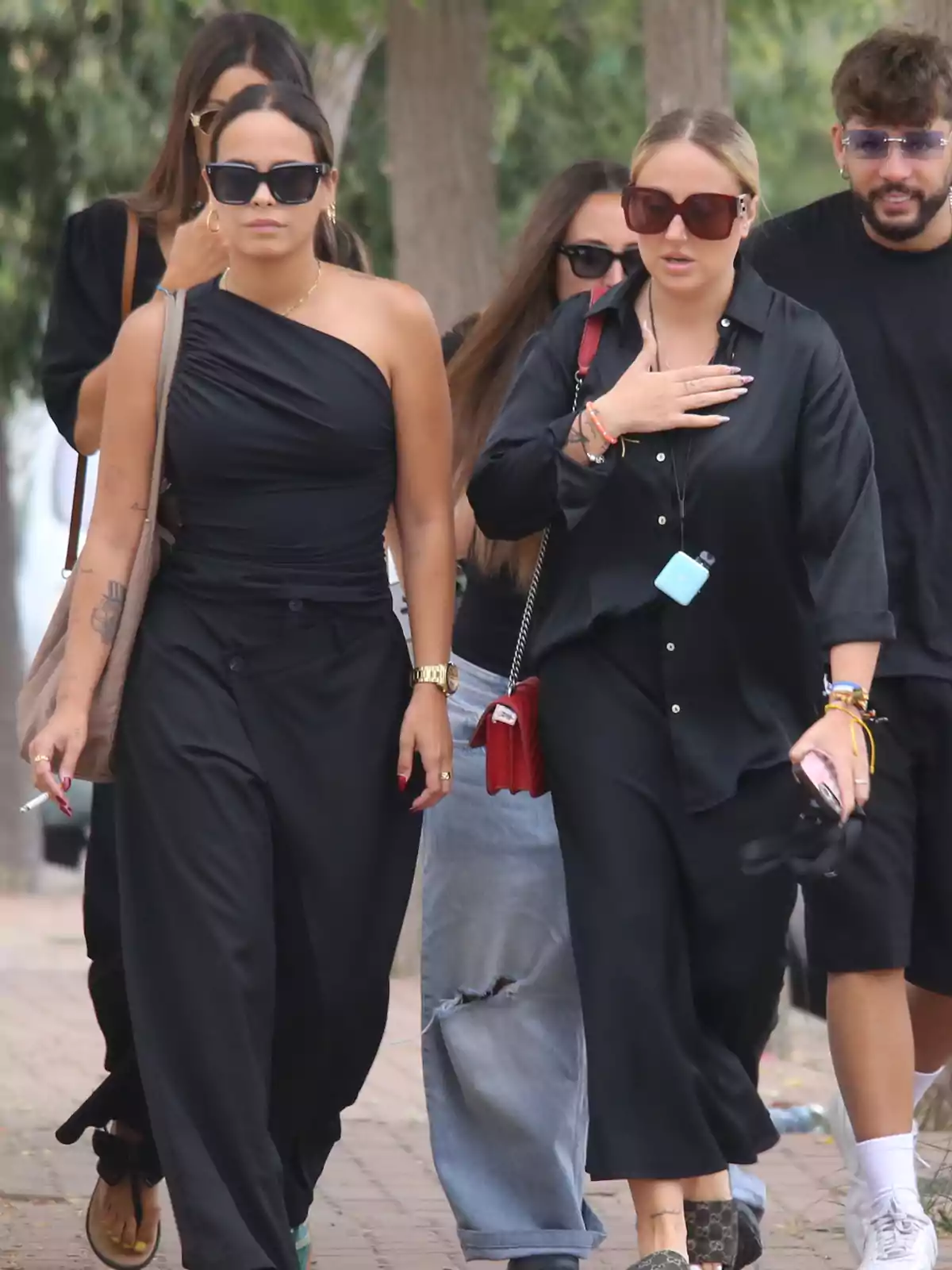 Grupo de personas caminando por la calle, la mayoría lleva gafas de sol y ropa oscura, al frente dos mujeres destacan por su vestimenta elegante y actitud seria Grupo de personas caminando por la calle, la mayoría lleva gafas de sol y ropa oscura, al frente dos mujeres destacan por su vestimenta elegante y actitud seria
