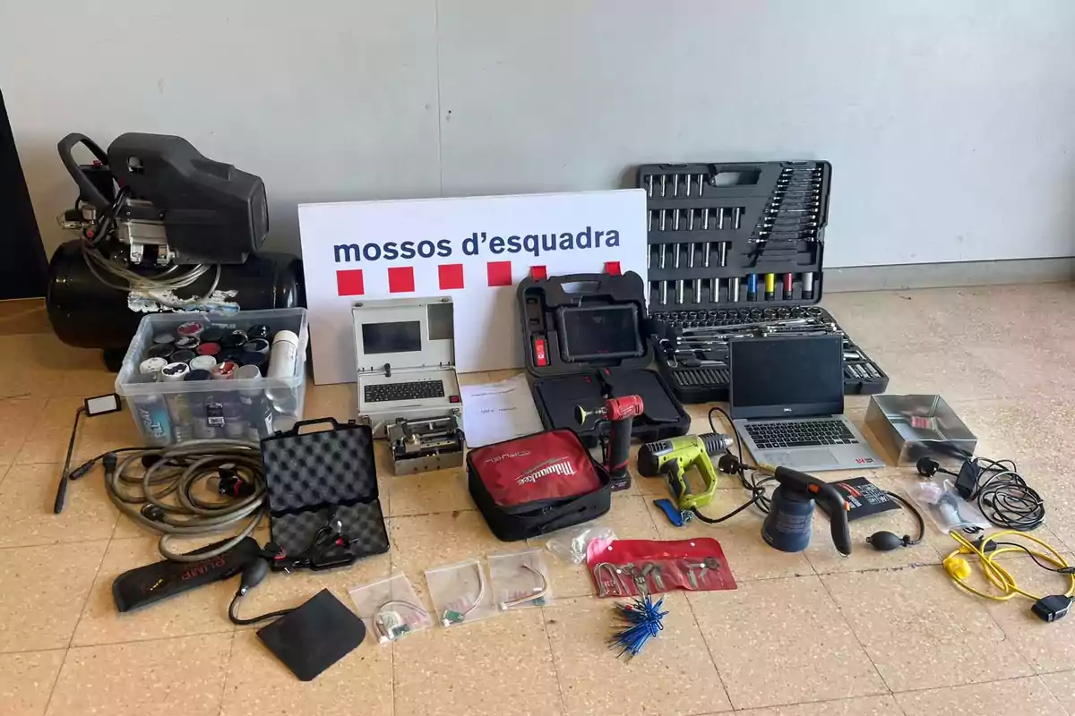 Varios equipos y herramientas como un compresor, ordenadores portátiles, maletines de herramientas, cables, sprays y dispositivos electrónicos están dispuestos en el suelo frente a un cartel de los Mossos d'Esquadra Varios equipos y herramientas como un compresor, ordenadores portátiles, maletines de herramientas, cables, sprays y dispositivos electrónicos están dispuestos en el suelo frente a un cartel de los Mossos d'Esquadra