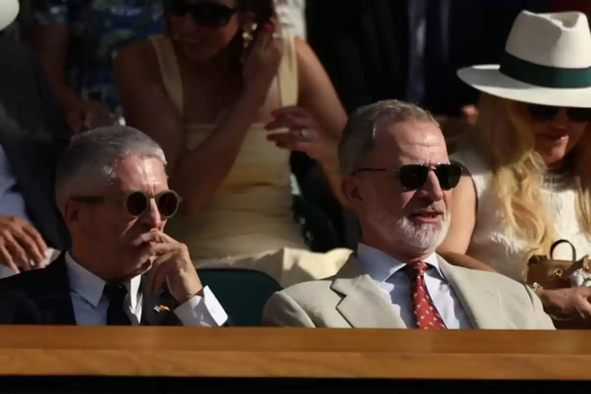 Dos hombres con gafas de sol sentados en un evento al aire libre, acompañados de más personas en el fondo Dos hombres con gafas de sol sentados en un evento al aire libre, acompañados de más personas en el fondo