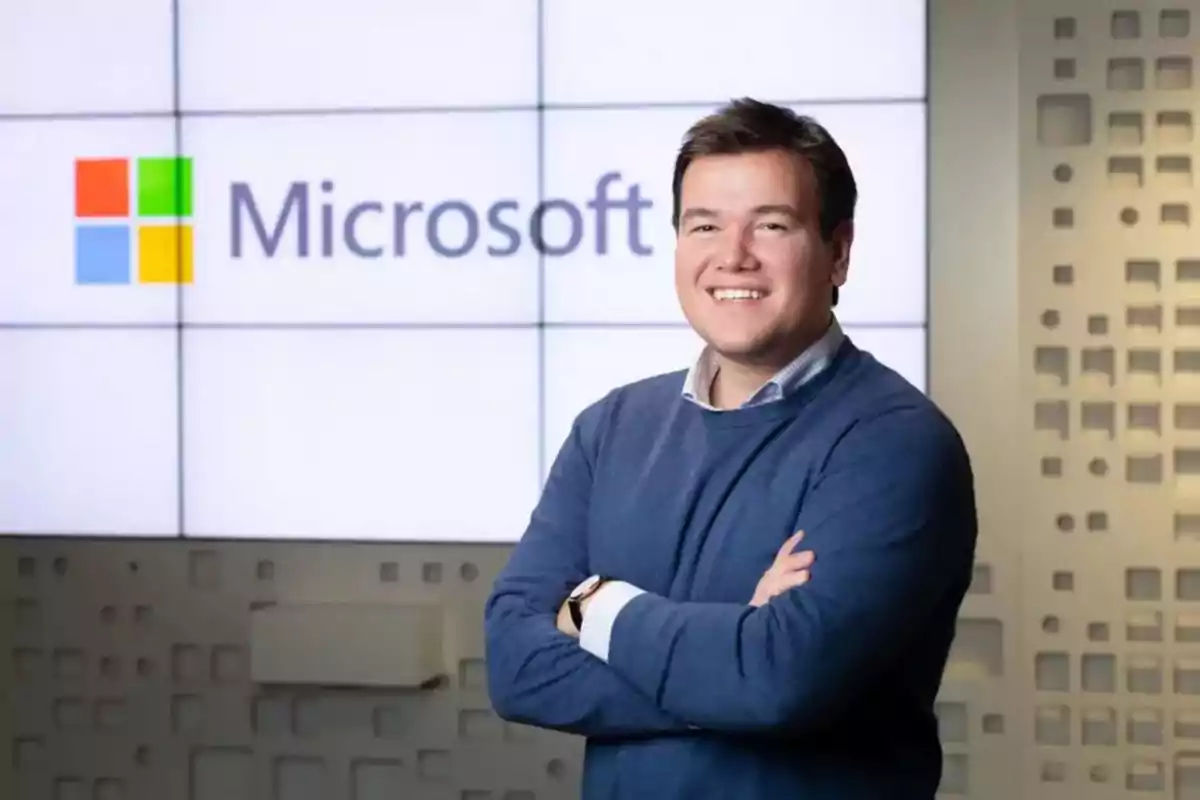 Hombre sonriente con suéter azul de pie frente a un logotipo de Microsoft en una pantalla grande
