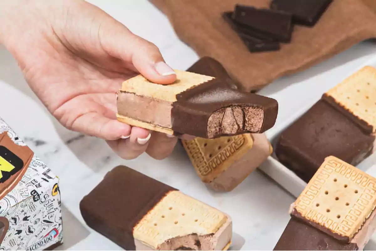 Una mano sostiene un sándwich de helado de chocolate cubierto parcialmente de chocolate, rodeado de otros sándwiches similares sobre una mesa Una mano sostiene un sándwich de helado de chocolate cubierto parcialmente de chocolate, rodeado de otros sándwiches similares sobre una mesa