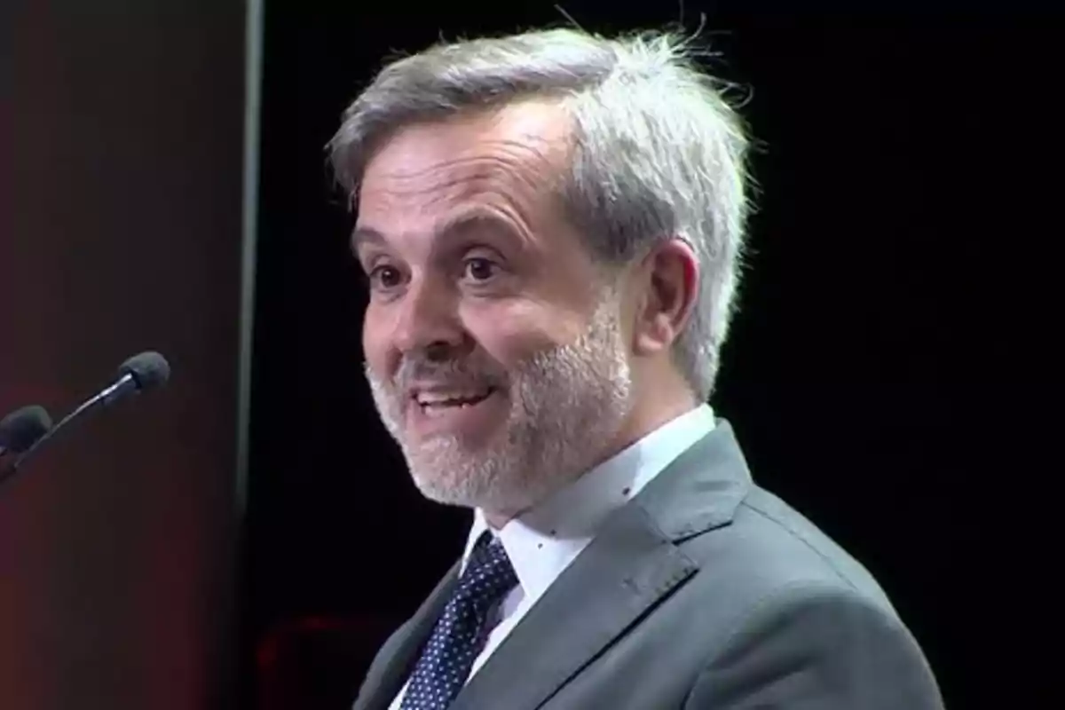 Hombre de cabello canoso y barba hablando sonriente frente a un micrófono con fondo oscuro Hombre de cabello canoso y barba hablando sonriente frente a un micrófono con fondo oscuro