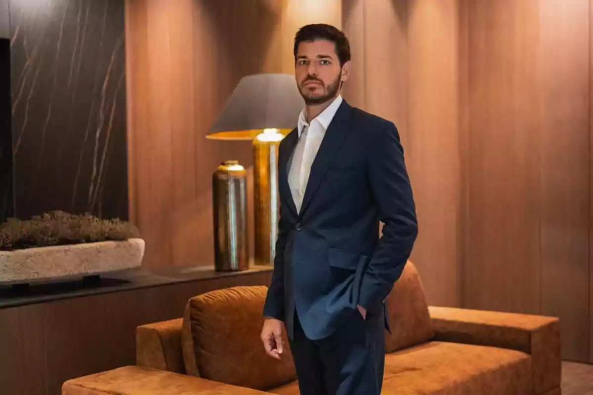 Hombre con barba y cabello oscuro vistiendo un traje azul marino y camisa blanca, de pie en una sala elegante con sofá marrón, lámpara dorada y paredes de madera