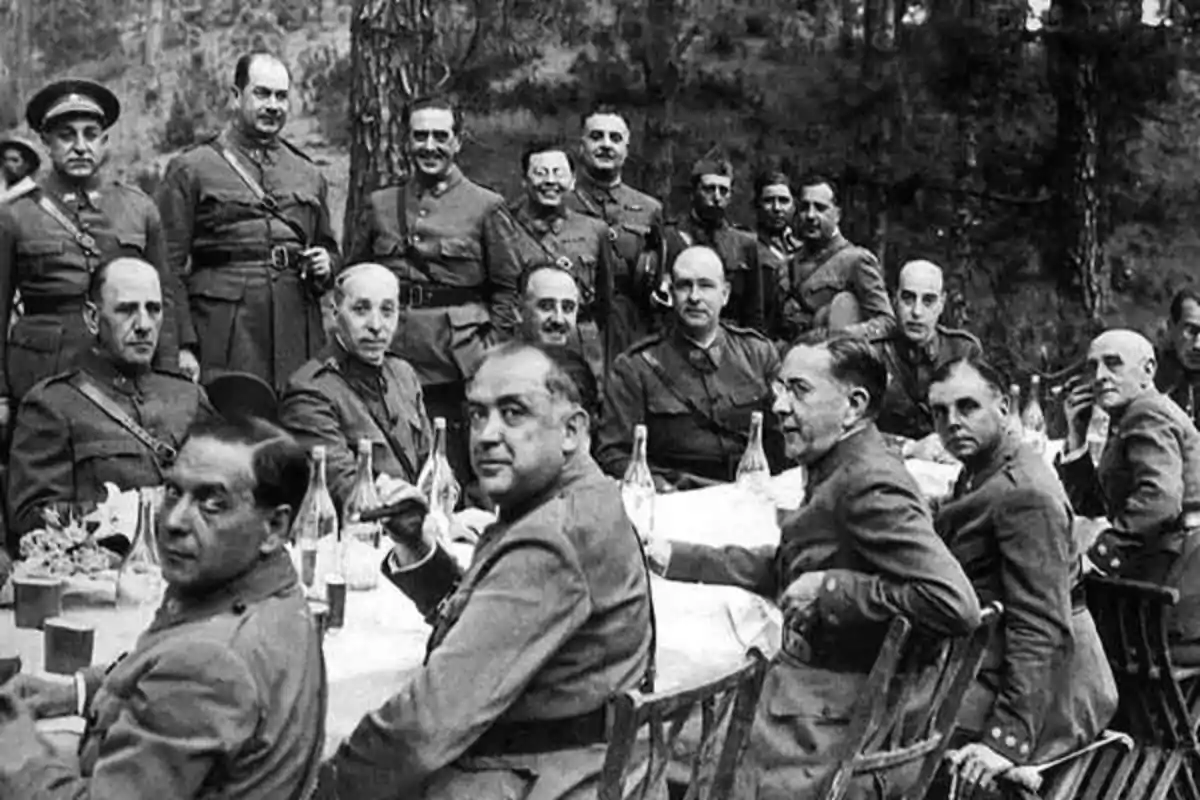 Un grupo de militares con uniformes antiguos está sentado y de pie alrededor de una mesa al aire libre en un entorno boscoso