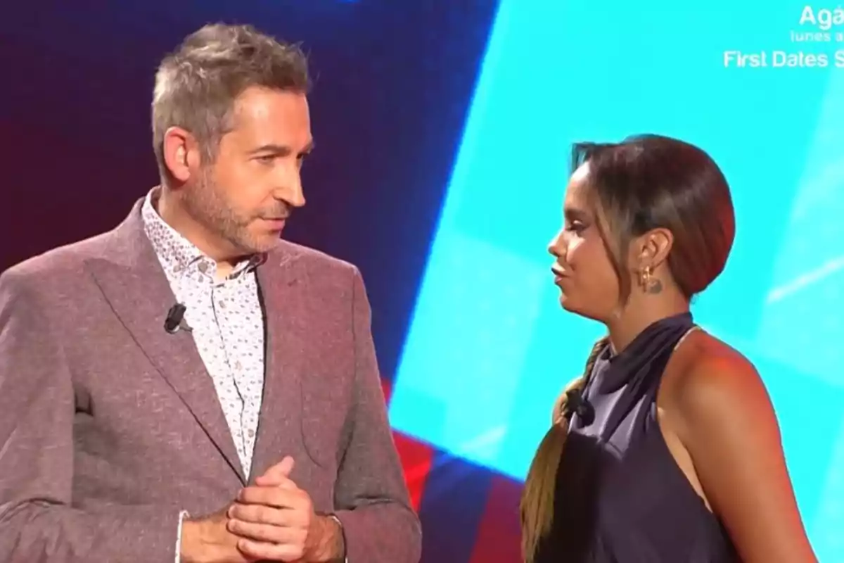 Frank Blanco con saco claro y Gloria Camila con vestido oscuro conversan en un set de televisión con fondo azul. Frank Blanco con saco claro y Gloria Camila con vestido oscuro conversan en un set de televisión con fondo azul.