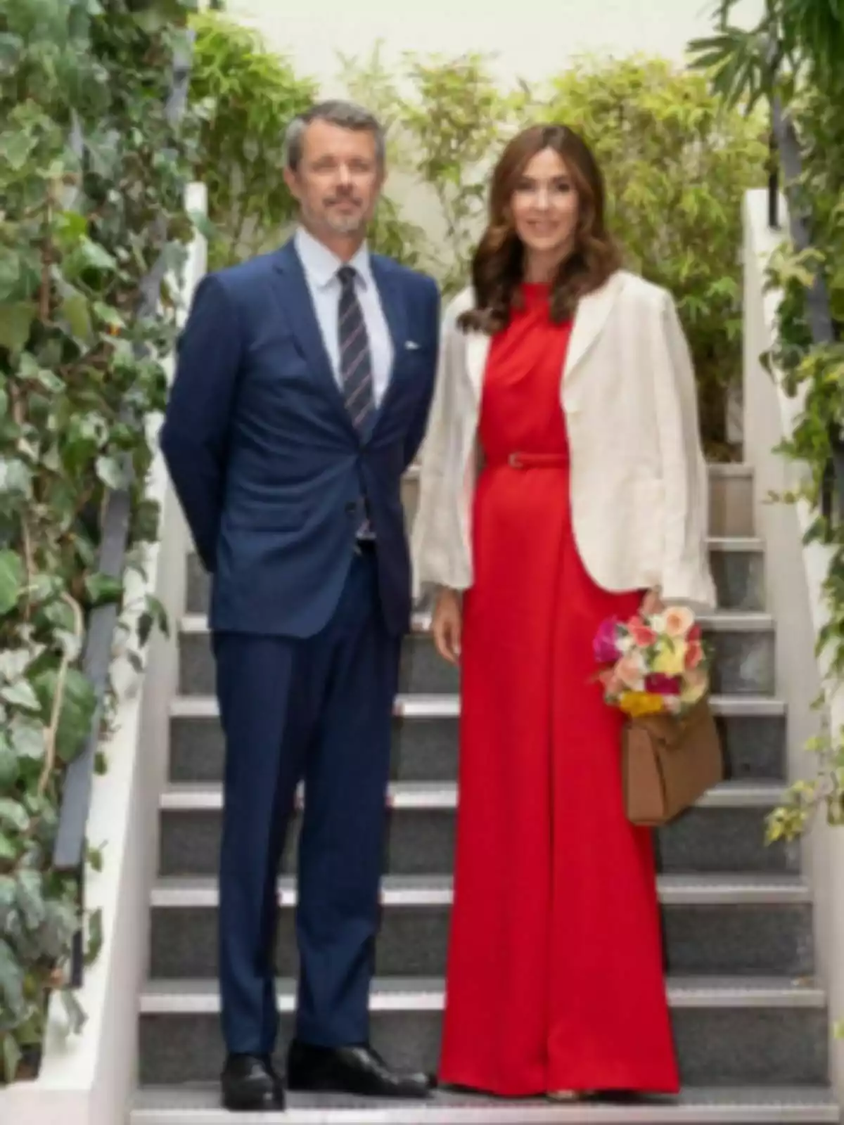 Pareja formada por Mary y Federico de Dinamarca posando en una escalera rodeada de plantas, el hombre lleva un traje azul y la mujer viste un conjunto rojo con chaqueta clara y sostiene un ramo de flores. Pareja formada por Mary y Federico de Dinamarca posando en una escalera rodeada de plantas, el hombre lleva un traje azul y la mujer viste un conjunto rojo con chaqueta clara y sostiene un ramo de flores.