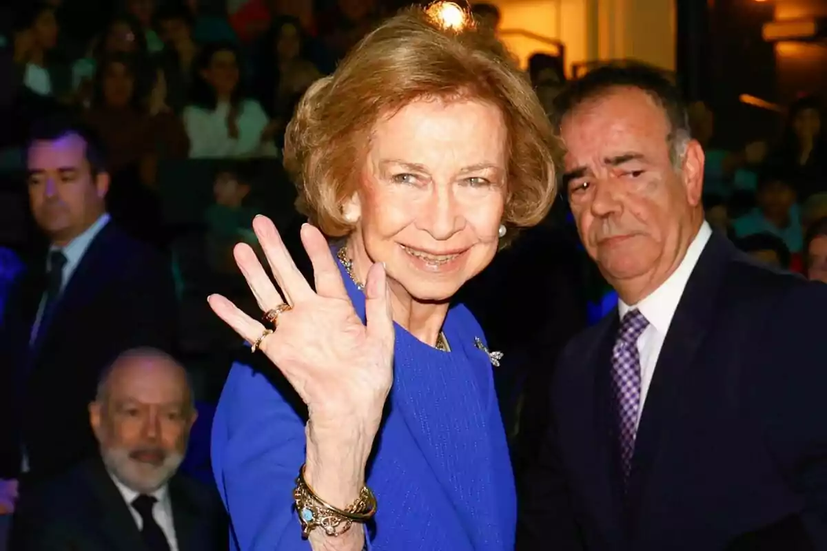 Mujer mayor con vestido azul saludando con la mano y sonriendo mientras varias personas la rodean en un evento formal Mujer mayor con vestido azul saludando con la mano y sonriendo mientras varias personas la rodean en un evento formal