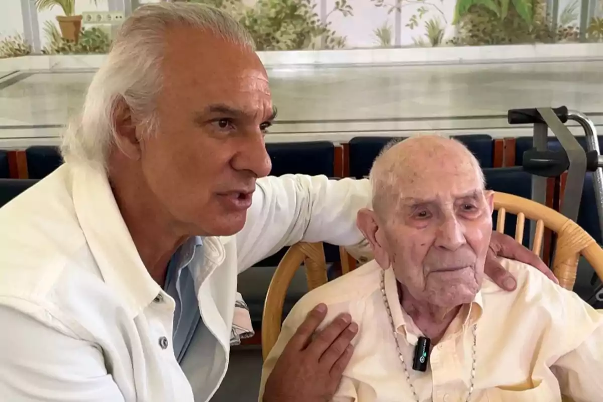 Dos hombres mayores sentados juntos, uno de ellos con el brazo sobre el hombro del otro, ambos mirando hacia el mismo lado en un ambiente interior iluminado