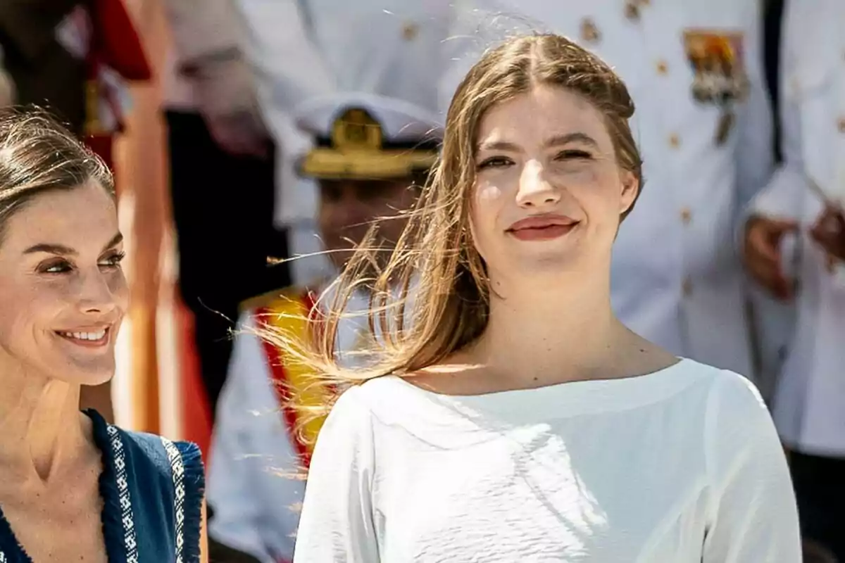 Two women smile outdoors during a formal event with uniformed people in the background. Two women smile outdoors during a formal event with uniformed people in the background.