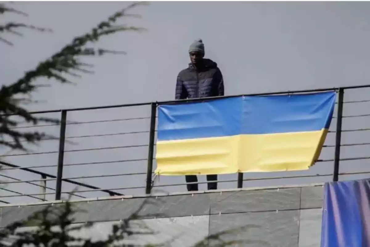 Persona con gorro y chaqueta oscura de pie detrás de una barandilla junto a una bandera de Ucrania