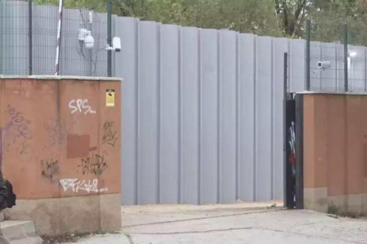 Muro alto de láminas metálicas grises detrás de una pared baja de color marrón con grafitis y cámaras de seguridad Muro alto de láminas metálicas grises detrás de una pared baja de color marrón con grafitis y cámaras de seguridad