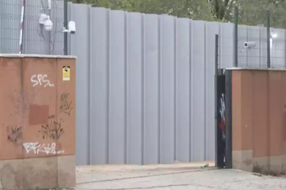 Muro de láminas metálicas grises detrás de una pared de ladrillo con grafitis y cámaras de seguridad