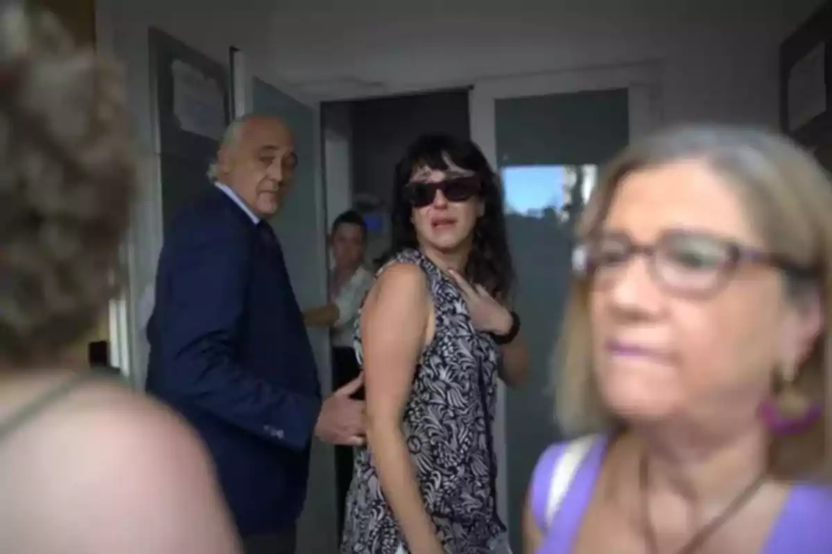Varias personas en un pasillo, una mujer con gafas oscuras parece emocionada o preocupada mientras un hombre de traje la acompaña y otras personas están desenfocadas en primer plano.