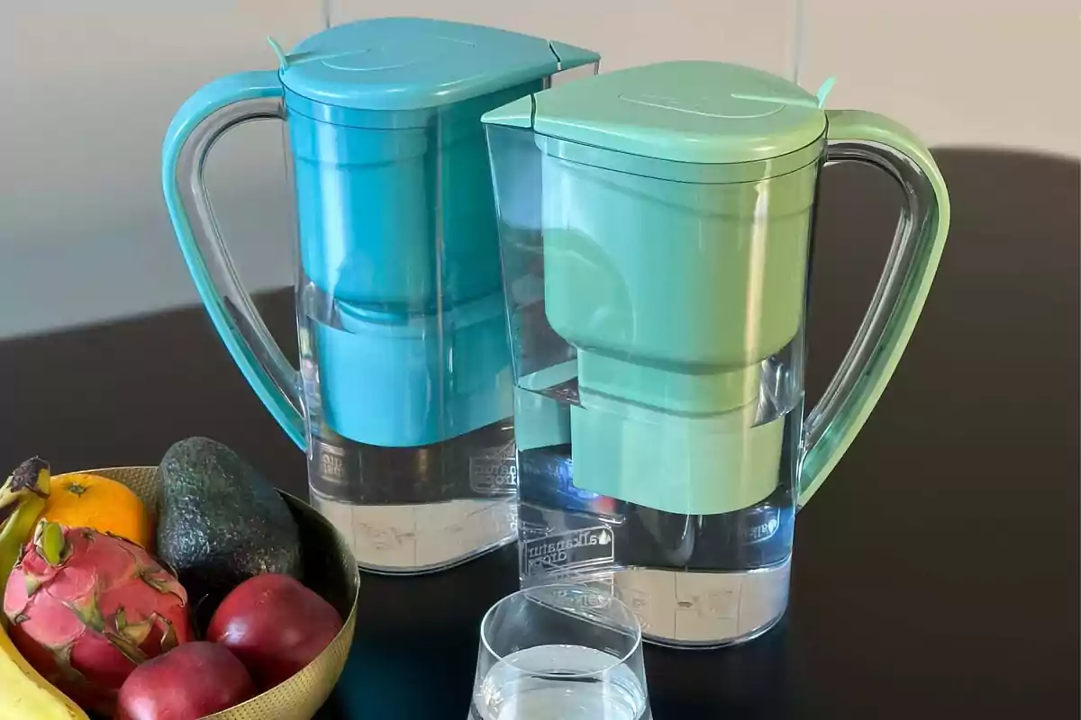 Dos jarras filtradoras de agua de colores azul y verde están sobre una mesa junto a un vaso de agua y un frutero con plátano, aguacate, pitaya, manzana y mandarina.