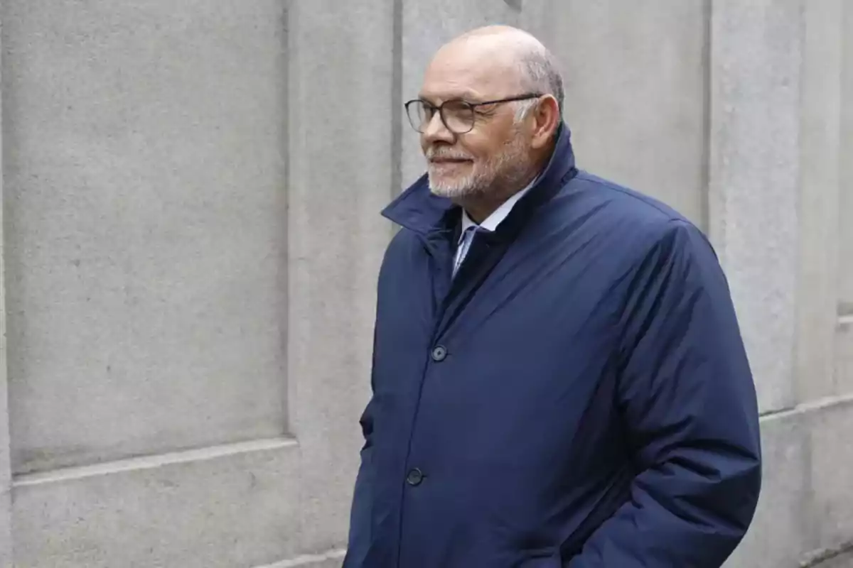 Hombre mayor con gafas y barba vestido con abrigo azul caminando junto a una pared de piedra Hombre mayor con gafas y barba vestido con abrigo azul caminando junto a una pared de piedra