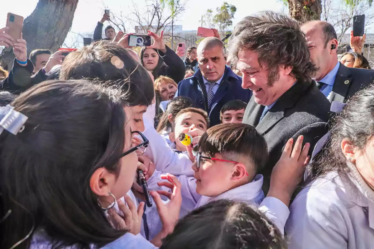 Un hombre sonriente rodeado de niños con guardapolvos blancos mientras varias personas toman fotos con sus teléfonos móviles en un ambiente al aire libre.