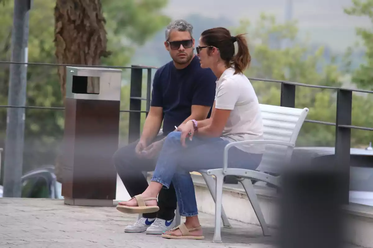 José Fernando y una mujer sentadas en una banca al aire libre conversan mientras usan gafas de sol rodeados de árboles y un basurero cercano José Fernando y una mujer sentadas en una banca al aire libre conversan mientras usan gafas de sol rodeados de árboles y un basurero cercano