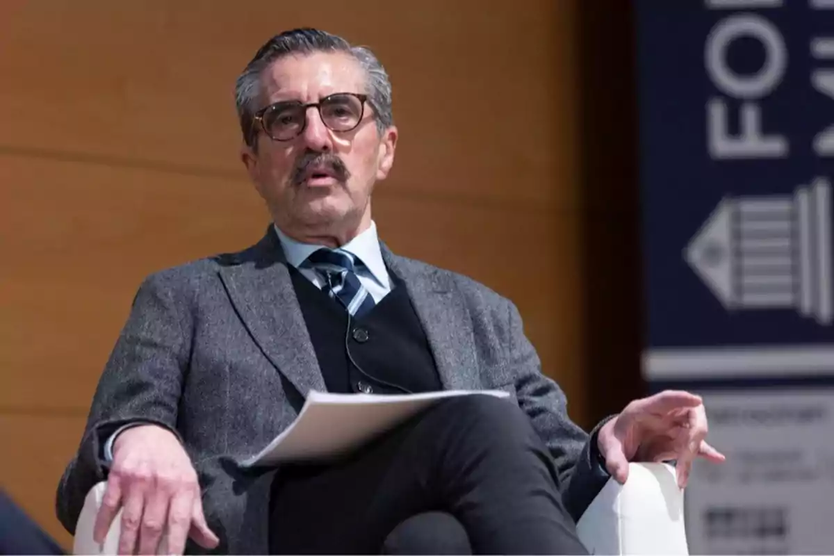 Hombre de mediana edad con gafas y bigote sentado en una silla vistiendo traje gris y corbata mientras sostiene unos papeles en la mano Hombre de mediana edad con gafas y bigote sentado en una silla vistiendo traje gris y corbata mientras sostiene unos papeles en la mano
