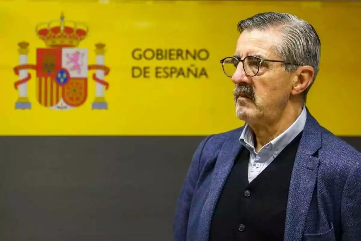Hombre de cabello canoso y gafas de pie frente a un fondo amarillo con el escudo y el texto Gobierno de España