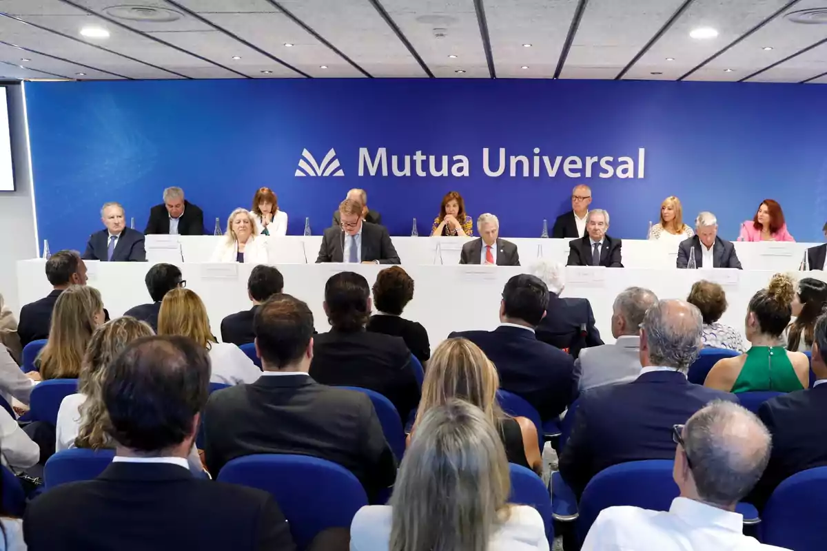 Personas sentadas en una sala de conferencias durante una reunión de Mutua Universal, con un panel de ponentes al frente y público asistente de espaldas.