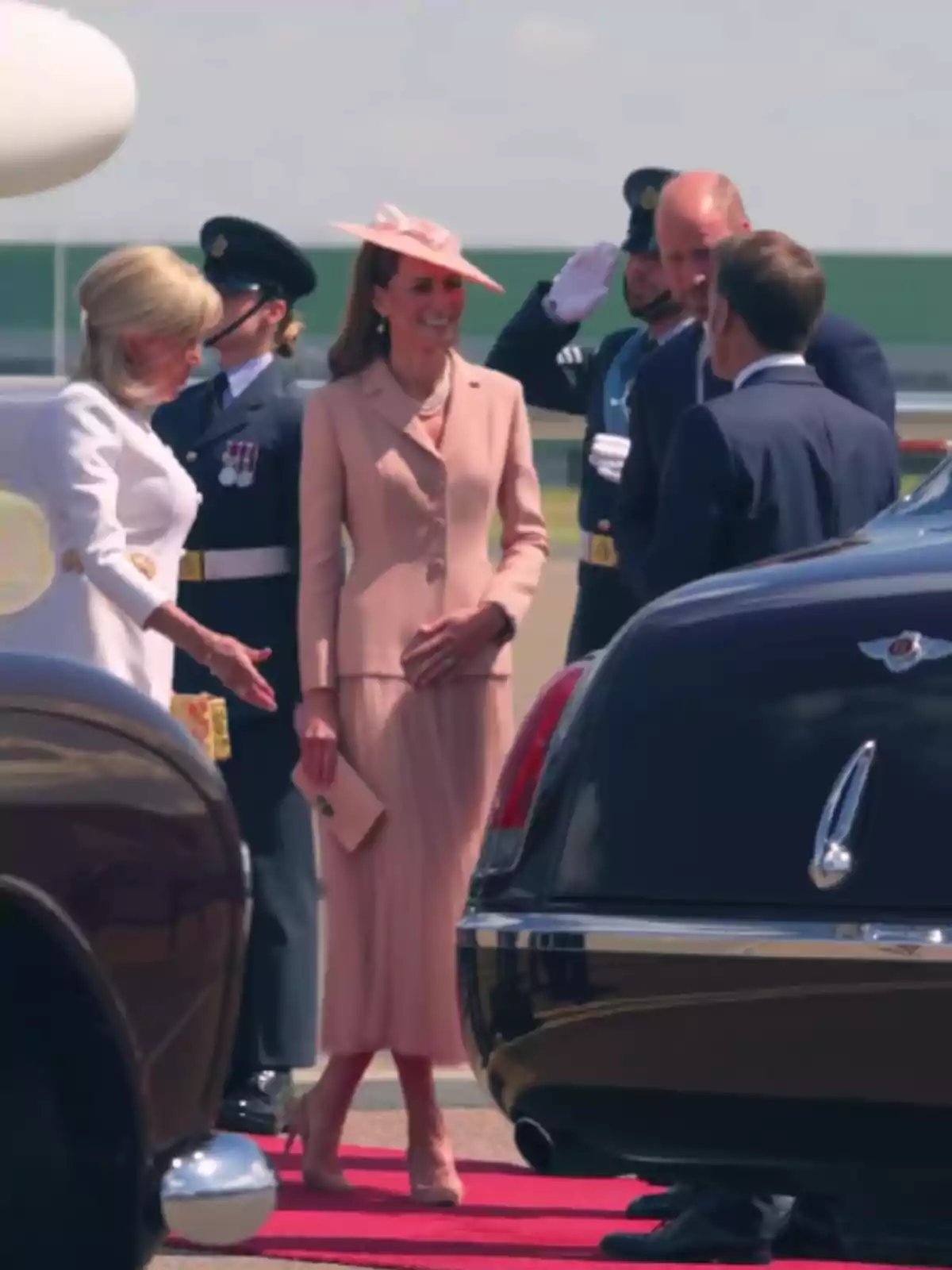 Several elegantly dressed people greet each other next to luxury cars on a red carpet at a formal outdoor event.